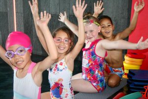 Smiling children in colorful swimwear and goggles joyfully raising their hands, highlighting the fun and inclusive atmosphere of Love to Swim's small group swimming lessons in East London.