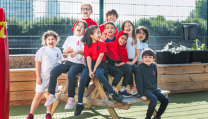 A group of cheerful young students in uniform sitting and standing on a wooden bench in an outdoor play area at a prep school in East London, enjoying a sunny day.
