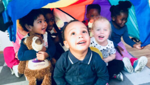 Young children take part in Kidslingo French and Spanish Classes for kids in East London- boys and girls of mixed ethnicities sit happily under a rainbow parachute. One boy is holding a teddy.