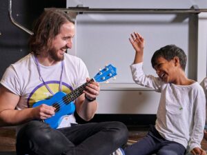 A man smiles as he sits teaching a a young boy the ukulele. February Half term is all about love and kIndness at the Strings Club