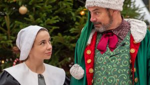 Two performers from Horrible Christmas stand in front of a festive Christmas tree. One is dressed in traditional historical attire with a white bonnet, while the other wears a vibrant, holiday-themed costume featuring a fur-trimmed green coat and festive details, capturing the playful spirit of the show.