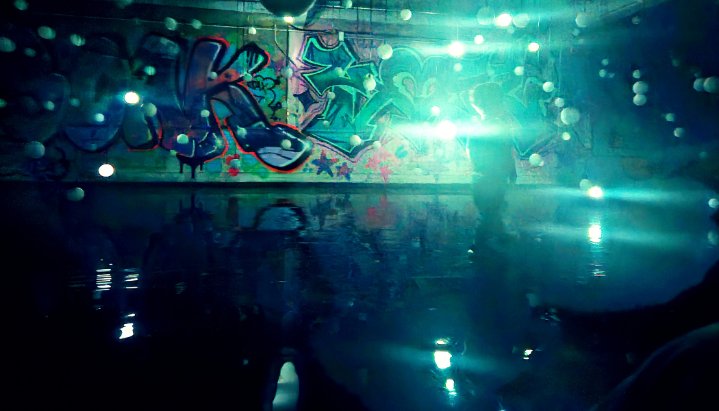 The image from East End Kids depicts a stunning scene from Lost in Light, blending urban art with immersive lighting. A graffiti-covered wall forms the backdrop, illuminated by glowing orbs suspended in mid-air. The room is filled with a reflective surface, enhancing the surreal atmosphere as light scatters across the space. A silhouette of a child interacting with the environment adds a human element, inviting viewers to imagine themselves immersed in this dynamic, otherworldly experience.
