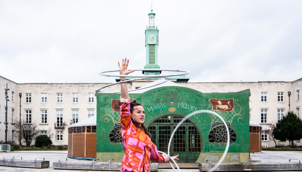 Performer spinning multiple hula hoops in vibrant attire in front of a green 'Magic Mirrors' structure at the Revel Puck Circus Winter Festival, with an art deco clock tower and historic building in the background
