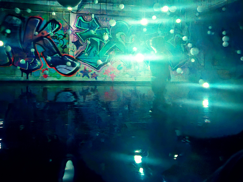 The image from East End Kids depicts a stunning scene from Lost in Light, blending urban art with immersive lighting. A graffiti-covered wall forms the backdrop, illuminated by glowing orbs suspended in mid-air. The room is filled with a reflective surface, enhancing the surreal atmosphere as light scatters across the space. A silhouette of a child interacting with the environment adds a human element, inviting viewers to imagine themselves immersed in this dynamic, otherworldly experience.