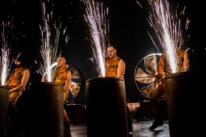 Production image from TRASH! with 4 men wearing high vis stand behind barrels shooting sparks high into the sky against a dark bacground their bodies illuminated by the sparks - a brilliant theatre experience for London Kids
