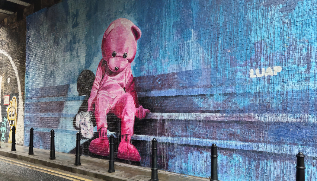 Giant pink teddy bear street art found in Shoreditch. Bear appears as a person in costume sat slumped on a bench in vivid pink against a blue background. East End kids rounds up things to do in Shoreditch with kids