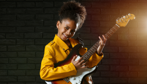 Black girl with curly hair in a bunch on top of her head smiles wearing a bright yellow jacket while holding an electric guitar - Strum stars Walthamstow half term holiday club