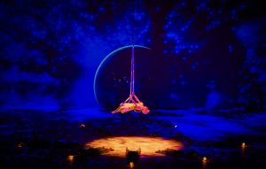 Review of The Little Prince – A breathtaking theatrical scene featuring an aerial performer suspended mid-air with flowing fabric, set against a cosmic backdrop of deep blue stars and a glowing planet. Dancers on the stage hold small illuminated orbs, enhancing the immersive and dreamlike atmosphere of this visually stunning production.