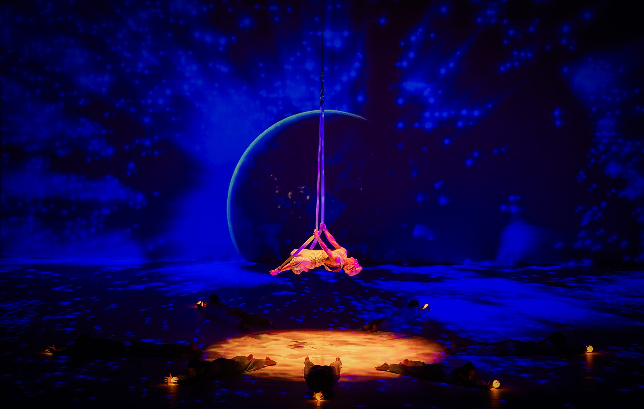 Review of The Little Prince – A breathtaking theatrical scene featuring an aerial performer suspended mid-air with flowing fabric, set against a cosmic backdrop of deep blue stars and a glowing planet. Dancers on the stage hold small illuminated orbs, enhancing the immersive and dreamlike atmosphere of this visually stunning production.
