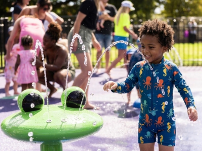 Little black boy plays in a frog shaped fountain at Clissold park splash pad. Parents mix with kids in soft focus in the background. It's a lovely sunny day and everything has a sunny sparkle.