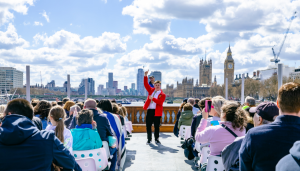 A lively group of people sit on the open-top river Terrible Thames tour boat cruising along the Thames in London. A tour guide in a red jacket stands at the front, gesturing as he speaks. In the background, the iconic skyline features Big Ben and the Houses of Parliament under a bright blue sky with scattered clouds.