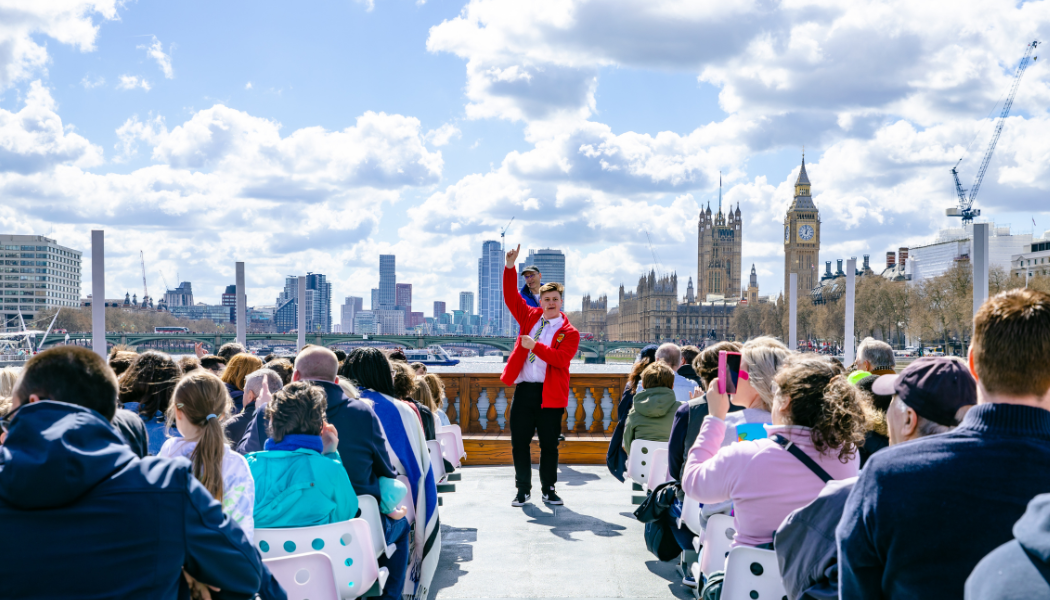 A lively group of people sit on the open-top river Terrible Thames tour boat cruising along the Thames in London. A tour guide in a red jacket stands at the front, gesturing as he speaks. In the background, the iconic skyline features Big Ben and the Houses of Parliament under a bright blue sky with scattered clouds.