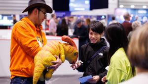 New Scientist Live brings the wonder of scientist to Excel London this October. A man holds dinosaur puppet out as a young boy holds out his hand to it's snout
