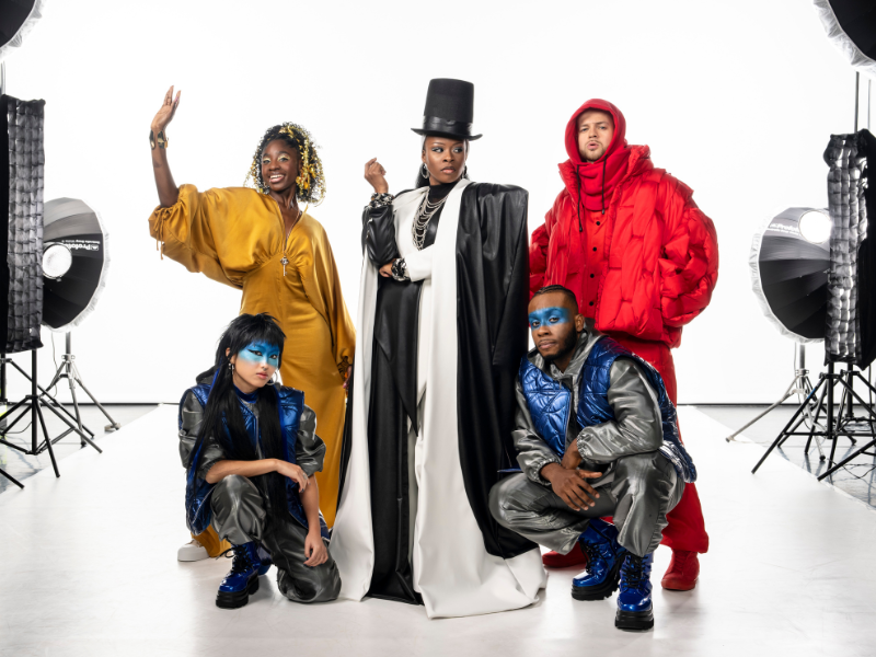 A group of five performers pose confidently in a brightly lit photography studio. At the centre stands a person in a dramatic black-and-white floor-length coat and tall black top hat, exuding a powerful, fashion-boss energy. To the left, a performer in a flowing golden dress lifts one arm with a joyful expression. To the right, another performer wears an oversized bright-red padded jacket and trousers. In front, two performers crouch in matching futuristic silver-and-blue outfits with vivid blue face paint and blue boots. Large studio lights and stands frame the white backdrop behind them. All performers from Ebony Scrooge coming to Sadler's Wells East this festive season