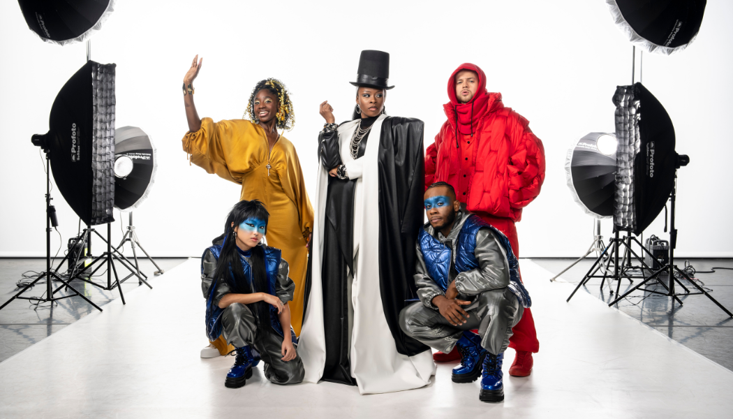 A group of five performers pose confidently in a brightly lit photography studio. At the centre stands a person in a dramatic black-and-white floor-length coat and tall black top hat, exuding a powerful, fashion-boss energy. To the left, a performer in a flowing golden dress lifts one arm with a joyful expression. To the right, another performer wears an oversized bright-red padded jacket and trousers. In front, two performers crouch in matching futuristic silver-and-blue outfits with vivid blue face paint and blue boots. Large studio lights and stands frame the white backdrop behind them. All performers from Ebony Scrooge coming to Sadler's Wells East this festive season
