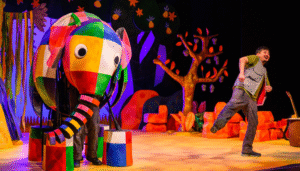 A brightly coloured stage shows a large patchwork Elmer the elephant puppet with bold squares of red, yellow, green, blue and purple, operated by performers whose legs are visible beneath the body. Beside Elmer, an actor in casual jungle-explorer clothing runs joyfully across the stage. The background features vibrant painted trees, plants and rocks in warm oranges and purples, creating a lively jungle setting. A perfect visual for families and kids in Walthamstow looking for a fun, colourful theatre adventure.