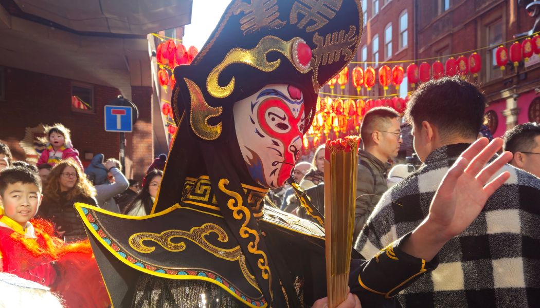 A performer in a traditional Chinese costume and painted mask walks through a crowded street during Lunar New Year celebrations, holding a fan, with rows of red lanterns hanging overhead and families watching in bright winter sunlight. With many places to celebrate Chinese New Year this February with kids in East London