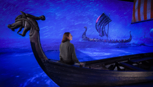 A woman sits in a Viking longboat bathed in cobalt blue light with projections of similar boats around her as part of a digital installation for the new VIkings: The Immersive Experience exhibition coming to East London this Spring