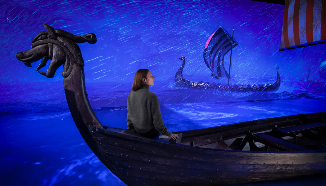 A woman sits in a Viking longboat bathed in cobalt blue light with projections of similar boats around her as part of a digital installation for the new VIkings: The Immersive Experience exhibition coming to East London this Spring
