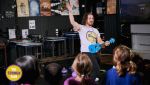 Children taking part in a music session at The Strings Club holiday camp, led by an instructor playing a ukulele.