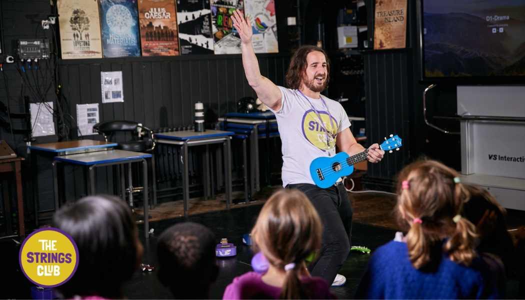 Children taking part in a music session at The Strings Club holiday camp, led by an instructor playing a ukulele.