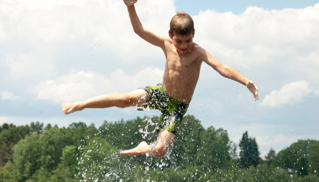 A young boy in green swim shorts leaps mid-air with arms and legs outstretched, mouth open in delight, against a backdrop of blue sky, white clouds and green treetops, with water droplets spraying around him